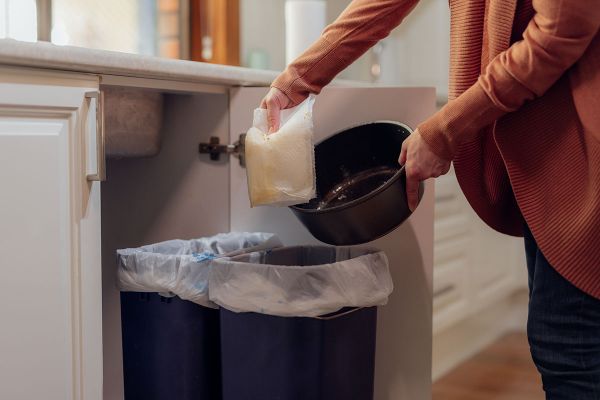 A woman’s hand holding a piece of fat-covered kitchen roll above a bag-lined waste bin pulled out from a kitchen cupboard. In the woman’s other hand is a black saucepan which has been used for cooking.